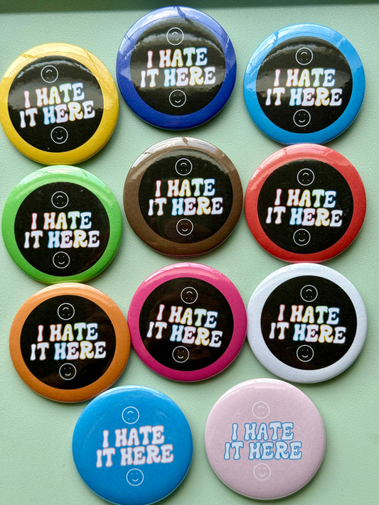 Assorted colorful buttons with 'I HATE IT HERE' text on a green background