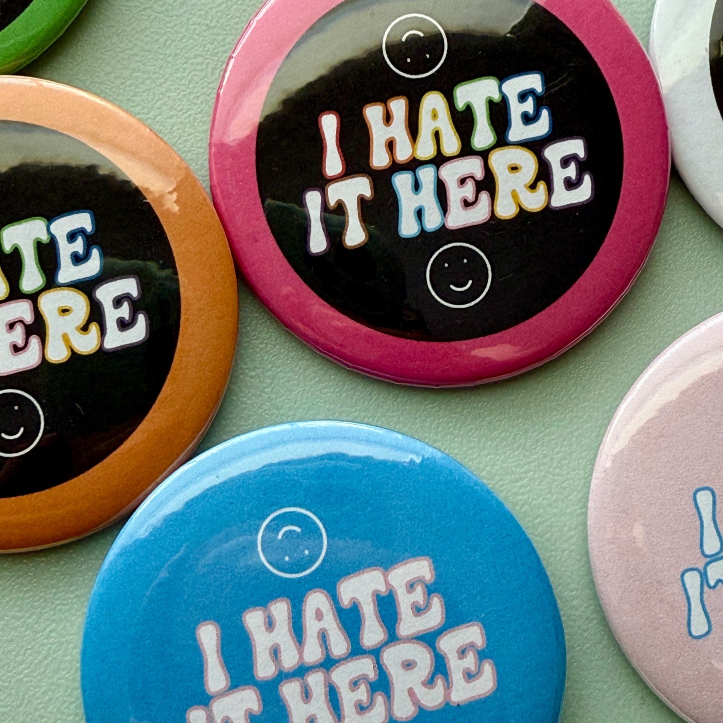 Collection of buttons with 'I HATE IT HERE' text on a teal background