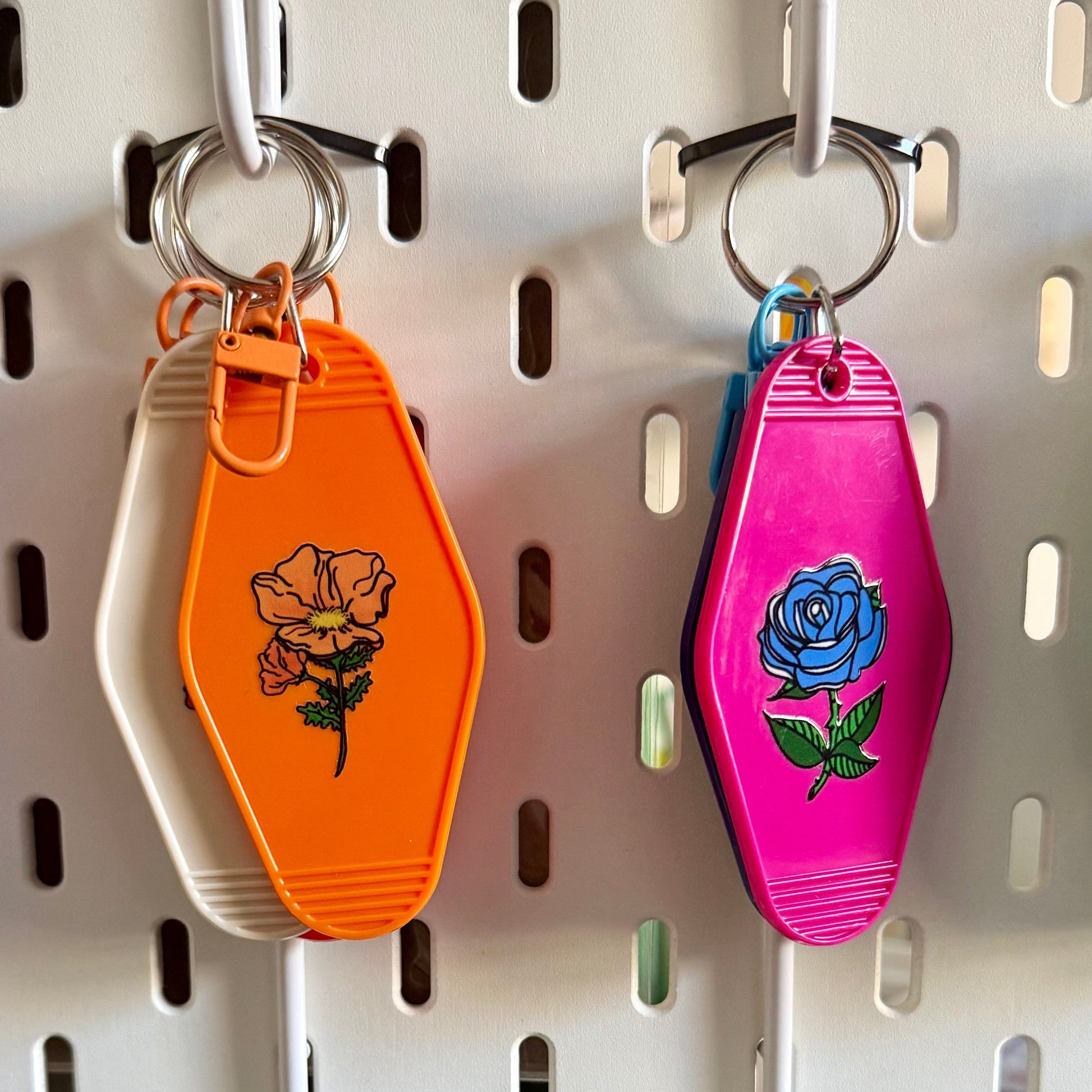 Four colorful coffin-shaped keychains hanging on a white pegboard. The keychains are orange, pink, light blue, and green, each decorated with a floral or heart design.