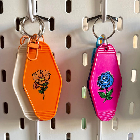 Four colorful coffin-shaped keychains hanging on a white pegboard. The keychains are orange, pink, light blue, and green, each decorated with a floral or heart design.