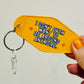 Yellow keychain with text 'I don't even want to be around anymore' held by a hand against a light gray background