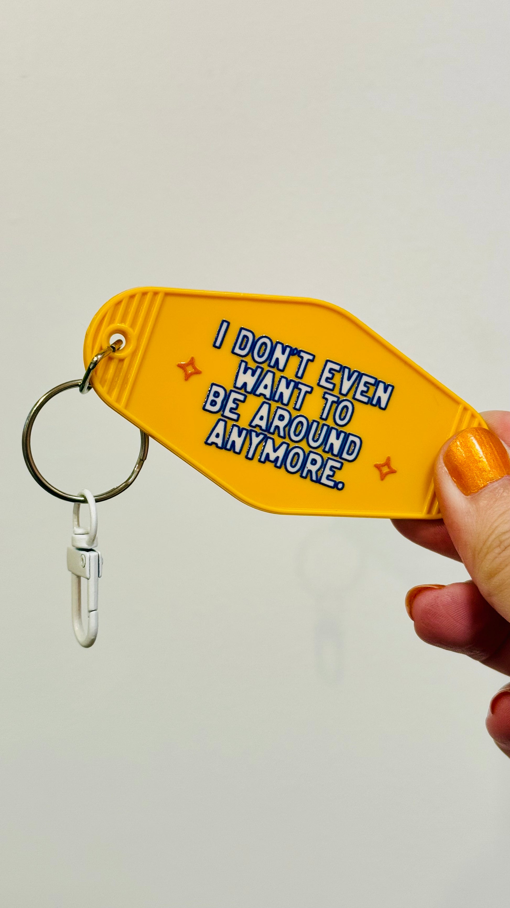 Yellow keychain with text 'I don't even want to be around anymore' held by a hand against a light gray background