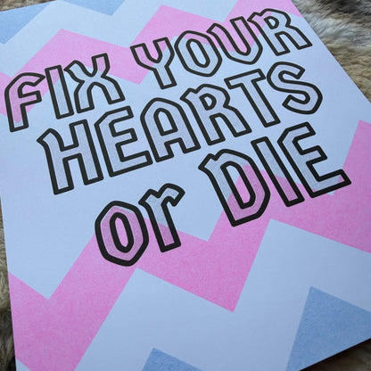 An art print with the text 'FIX YOUR HEARTS OR DIE' in bold black outline lettering over a pastel pink, light blue, and white geometric chevron background, resting on a textured brown surface.