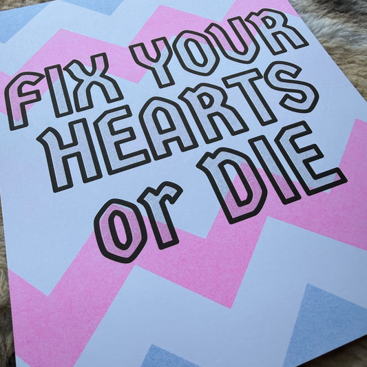 An art print with the text 'FIX YOUR HEARTS OR DIE' in bold black outline lettering over a pastel pink, light blue, and white geometric chevron background, resting on a textured brown surface.