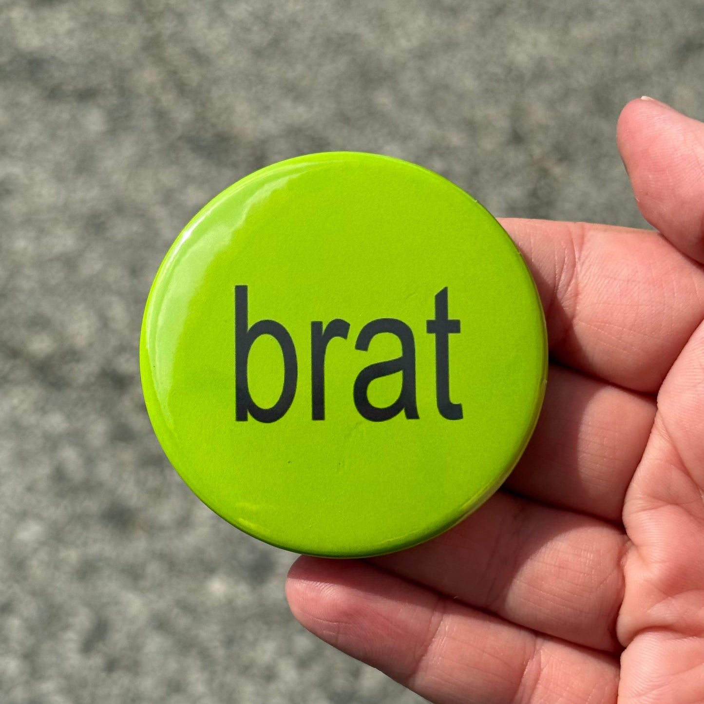 A hand holding a round lime green button badge with the word 'brat' printed in black lowercase letters, set against a blurred gray asphalt background.