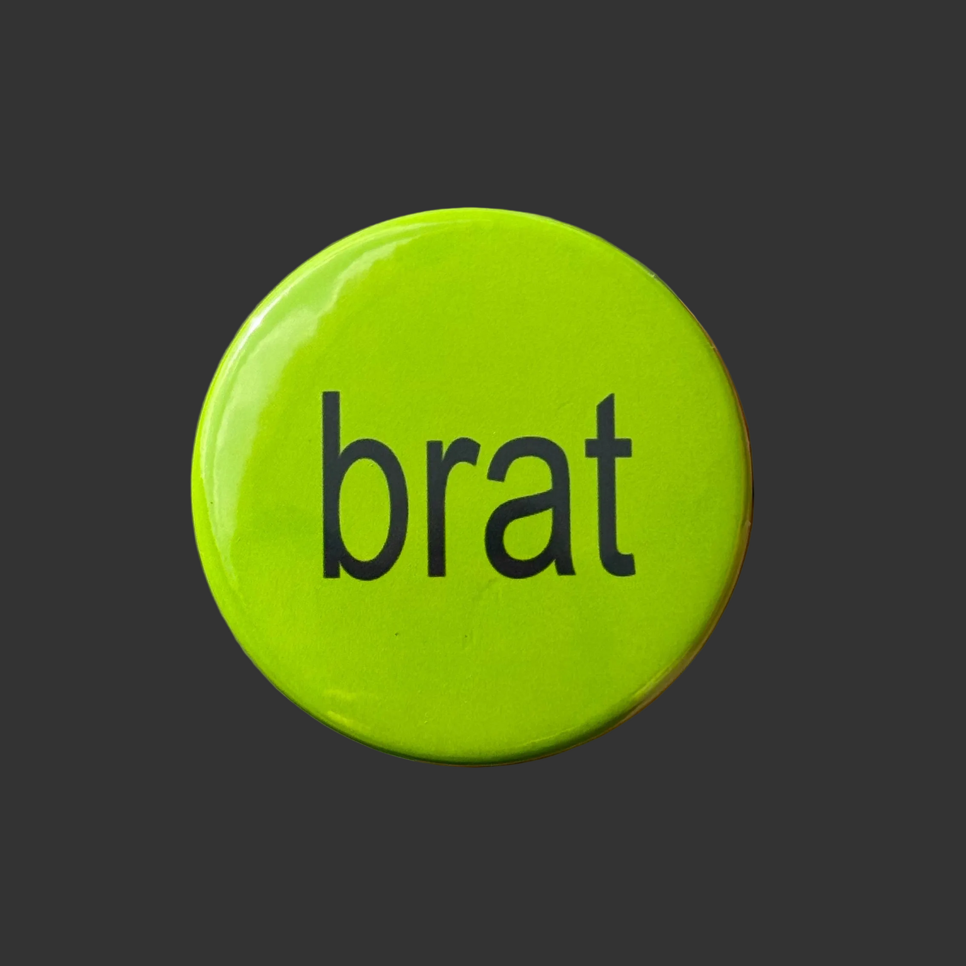 A round, glossy lime green button pin featuring the word 'brat' in a black lowercase sans-serif font, centered on a dark gray background.