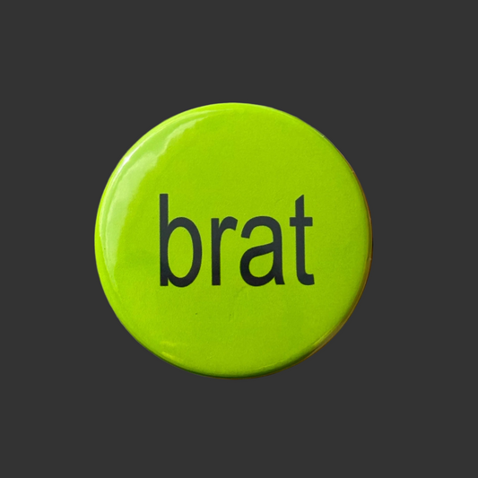 A round, glossy lime green button pin featuring the word 'brat' in a black lowercase sans-serif font, centered on a dark gray background.