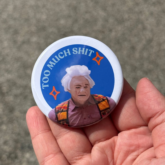 A hand holding a round button badge featuring a printed graphic of a person with the text 'TOO MUCH SHIT' on a blue background.