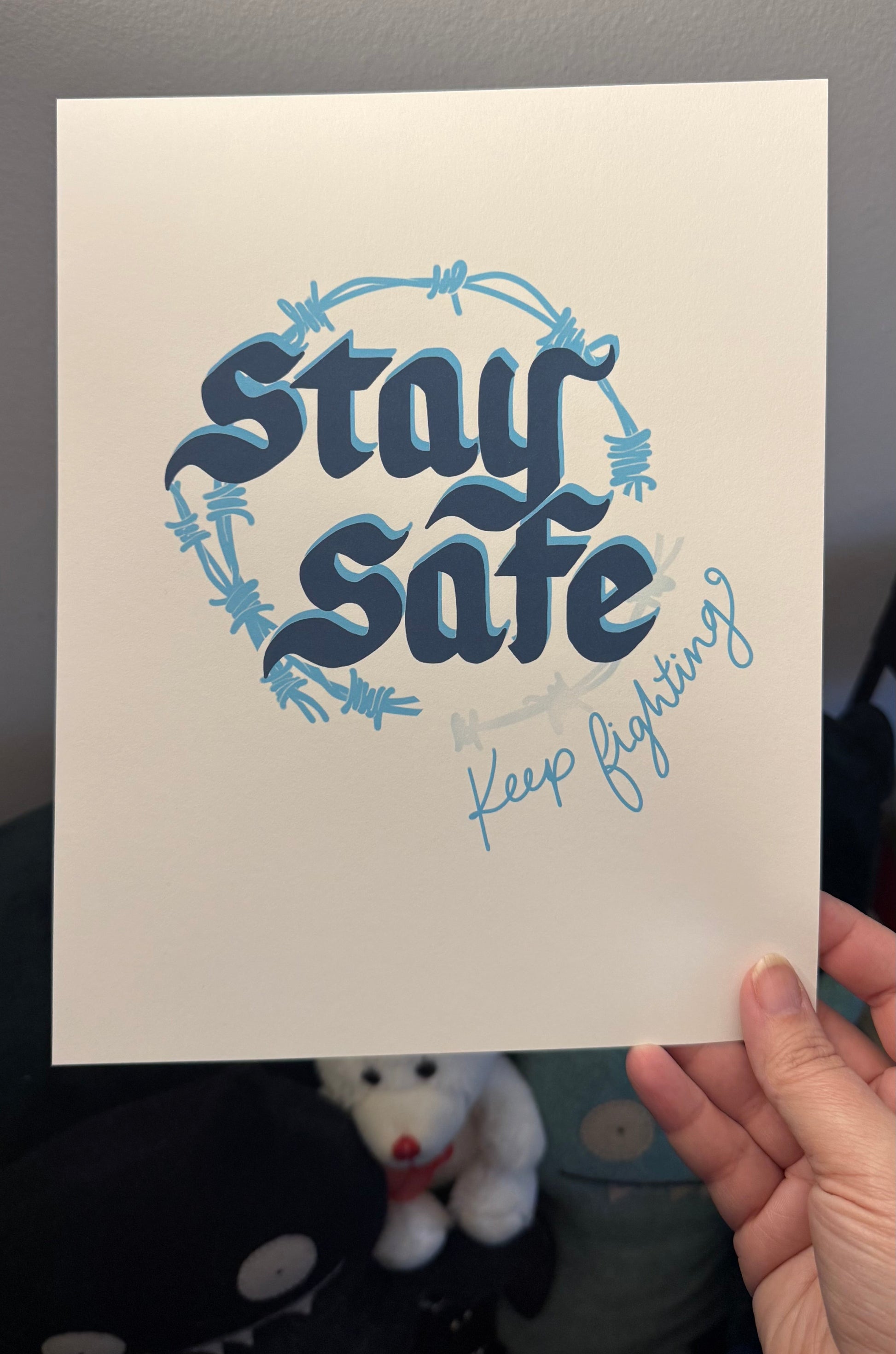 Hand holding a print with 'Stay Safe, Keep Fighting' text on a neutral background