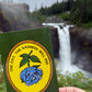 A hand holds a green card featuring a circular yellow emblem with a blue rose and the text 'ONE DAY, THE SADNESS WILL END' in front of a blurred waterfall landscape.