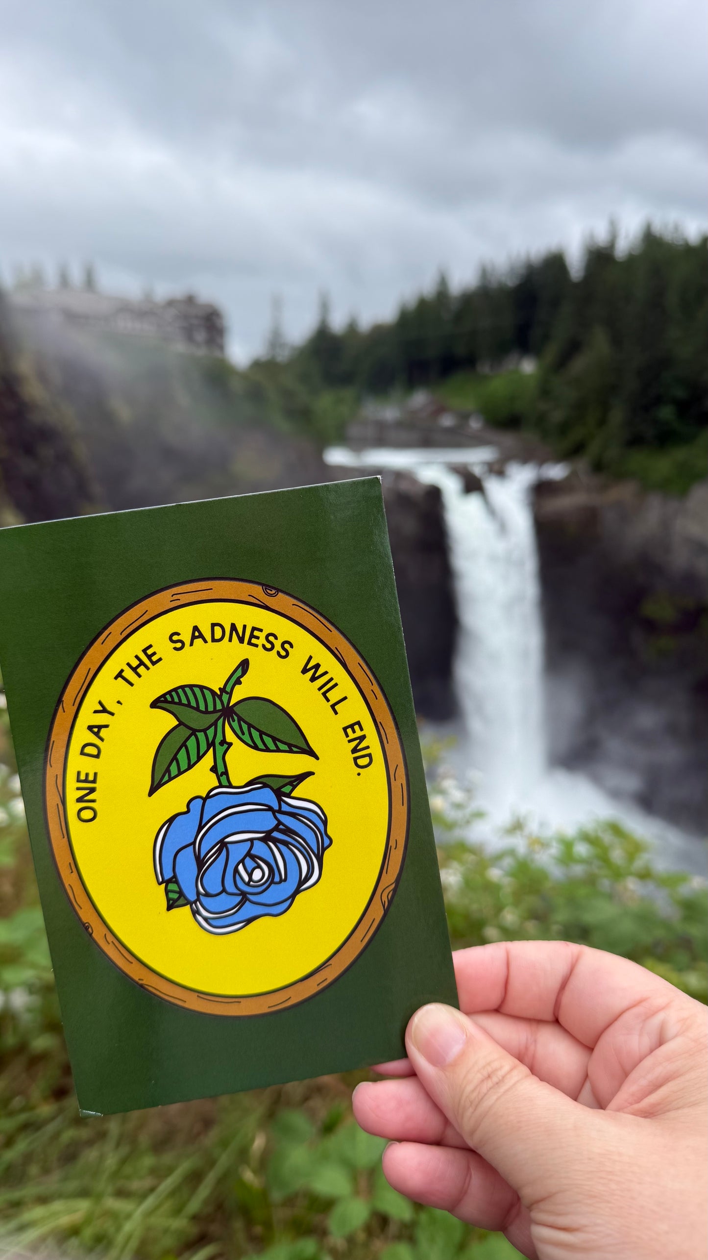A hand holds a green card featuring a circular yellow emblem with a blue rose and the text 'ONE DAY, THE SADNESS WILL END' in front of a blurred waterfall landscape.