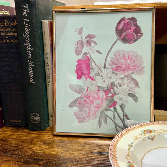 A framed vintage-style floral print featuring pink and mauve flowers on a white background, displayed on a wooden shelf next to books and a decorative plate.