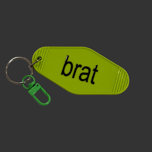 A lime green keychain tag with the word 'brat' printed in black lowercase letters, attached to a silver split ring and a green lobster clasp, shown on a dark gray background.